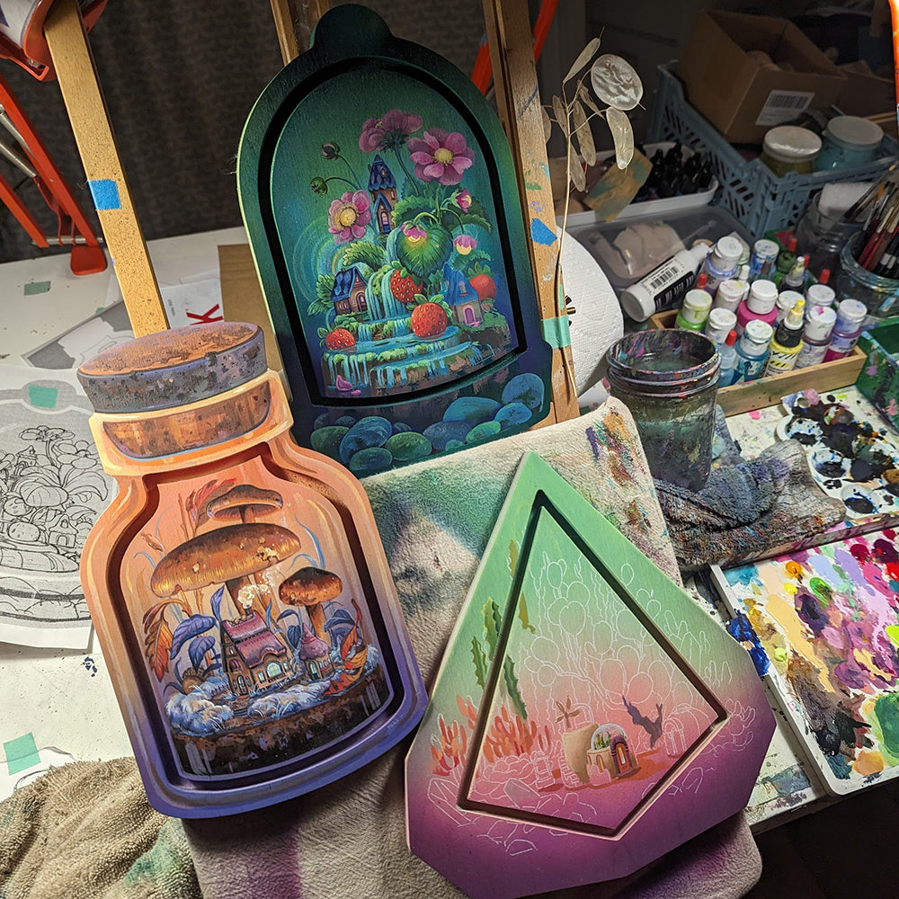 Painted bottle-shaped, arched and geometric panels amid brushes and paint containers on a studio table.