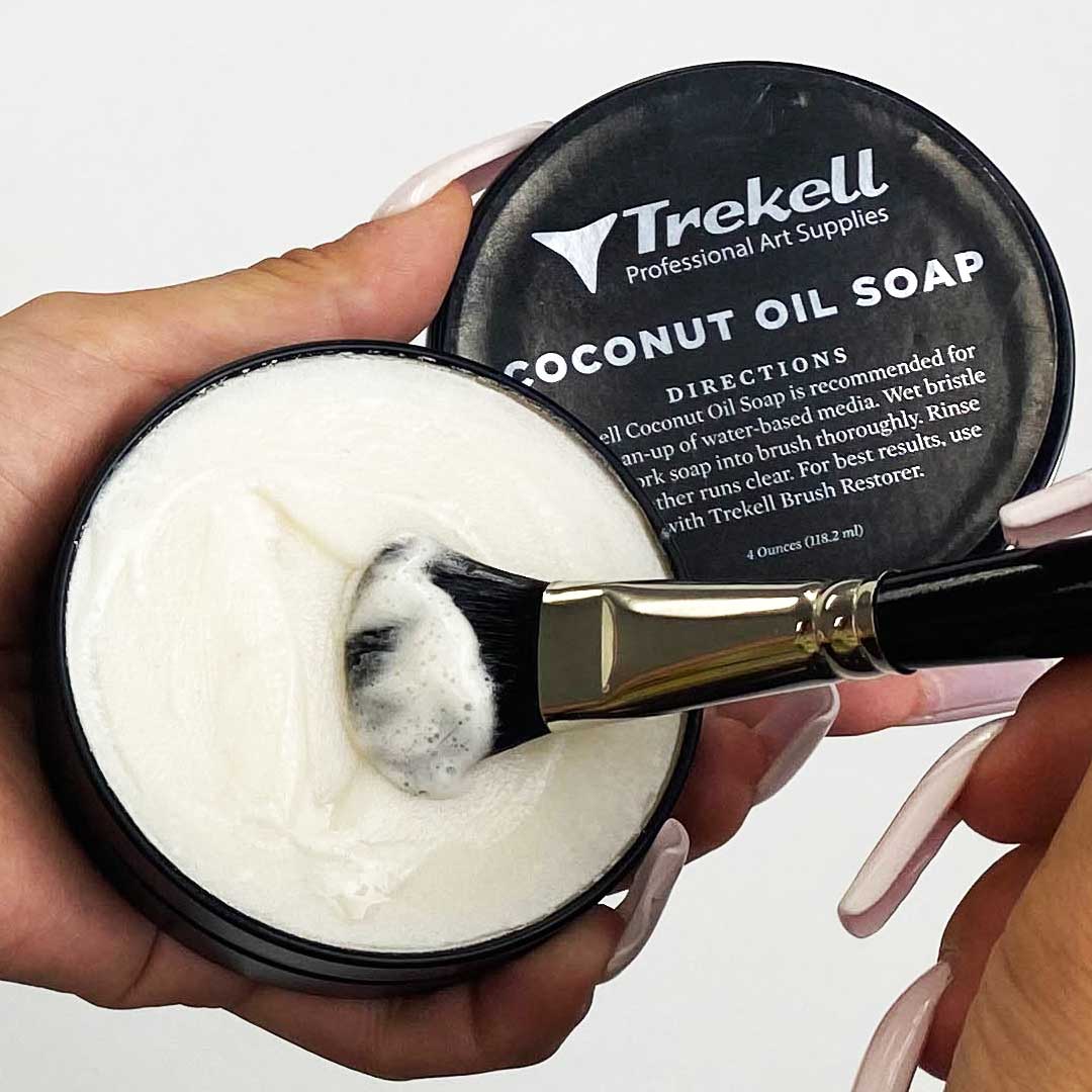Hands swirl a broad brush in white coconut oil soap inside an open black tin.