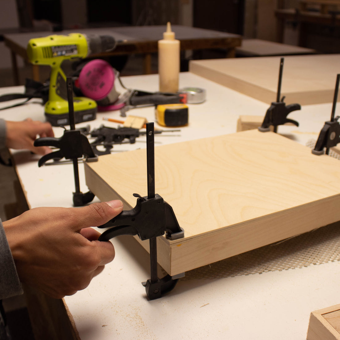 Hands clamp a rectangular wood panel on a workshop bench.
