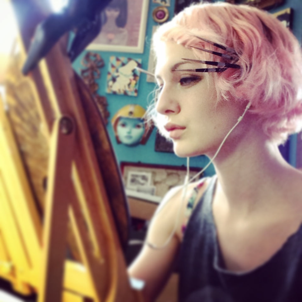 Pink-haired artist looks toward a painting at an easel.