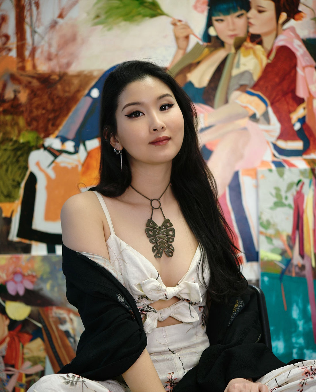 Long-haired woman sits before colorful paintings in a studio.