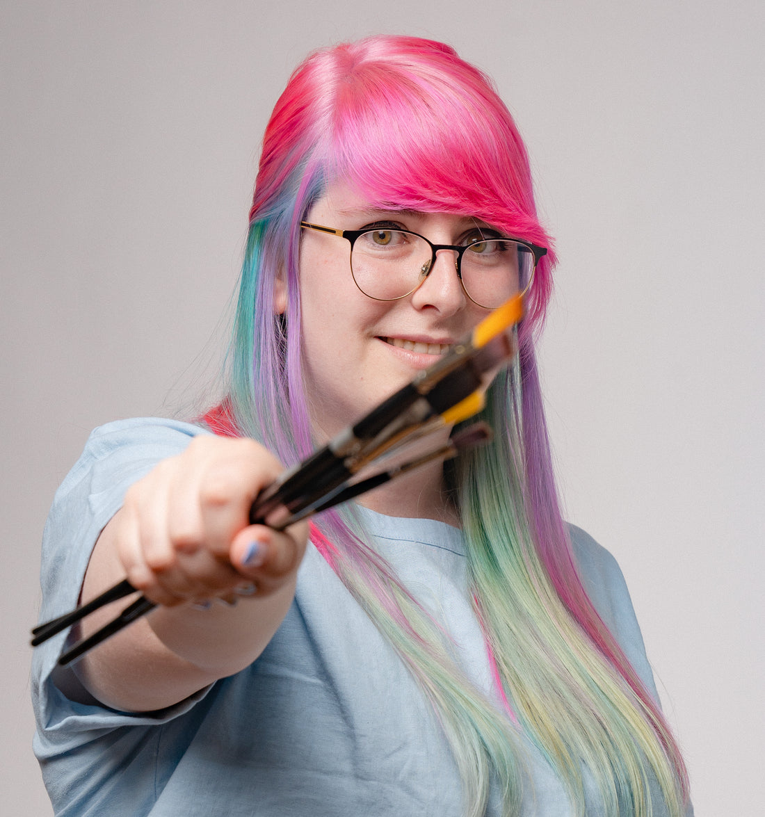 Smiling artist with pink-and-green hair and glasses holds brushes toward the camera.