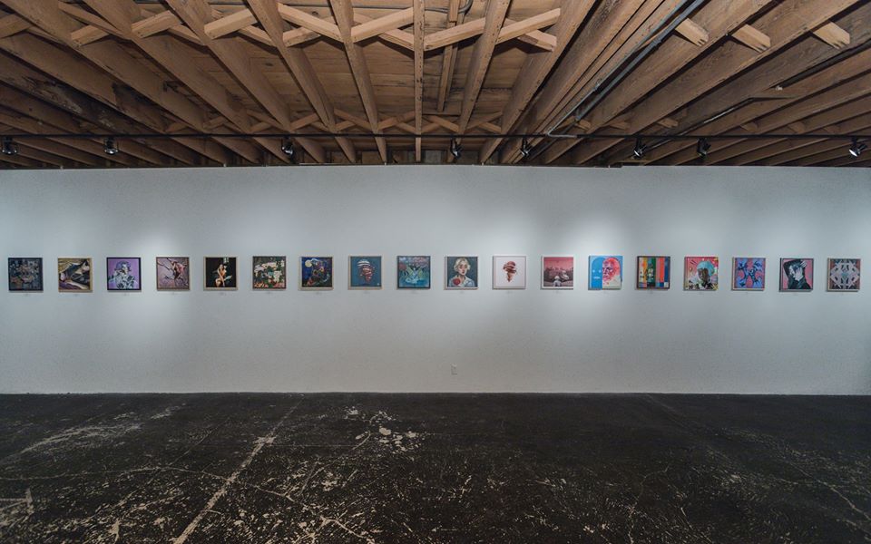Long row of small artworks in a gallery beneath exposed wooden ceiling beams.
