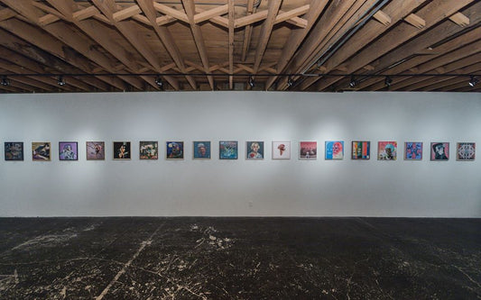 Long row of small artworks in a gallery beneath exposed wooden ceiling beams.