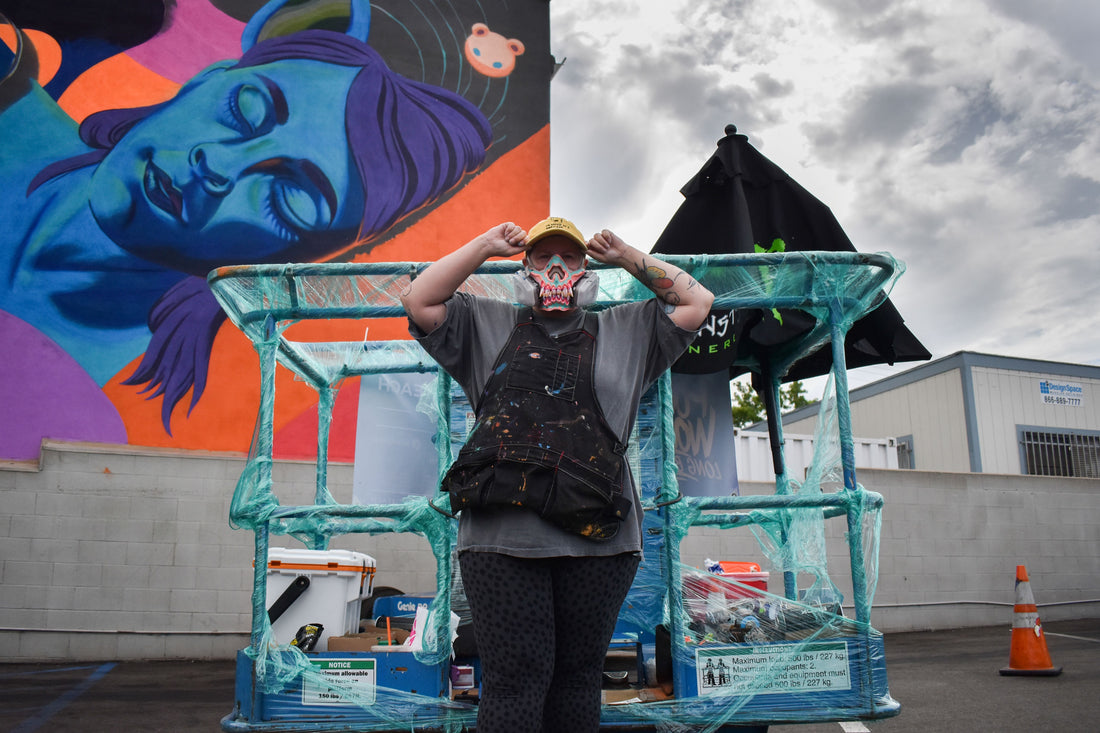 Artist stands beside a turquoise lift beneath a colorful outdoor mural.