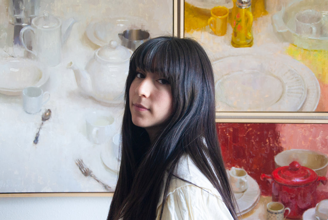 Dark-haired woman stands in front of large still-life paintings.