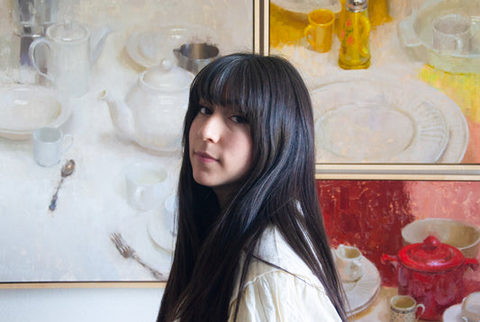 Dark-haired woman stands in front of large still-life paintings.