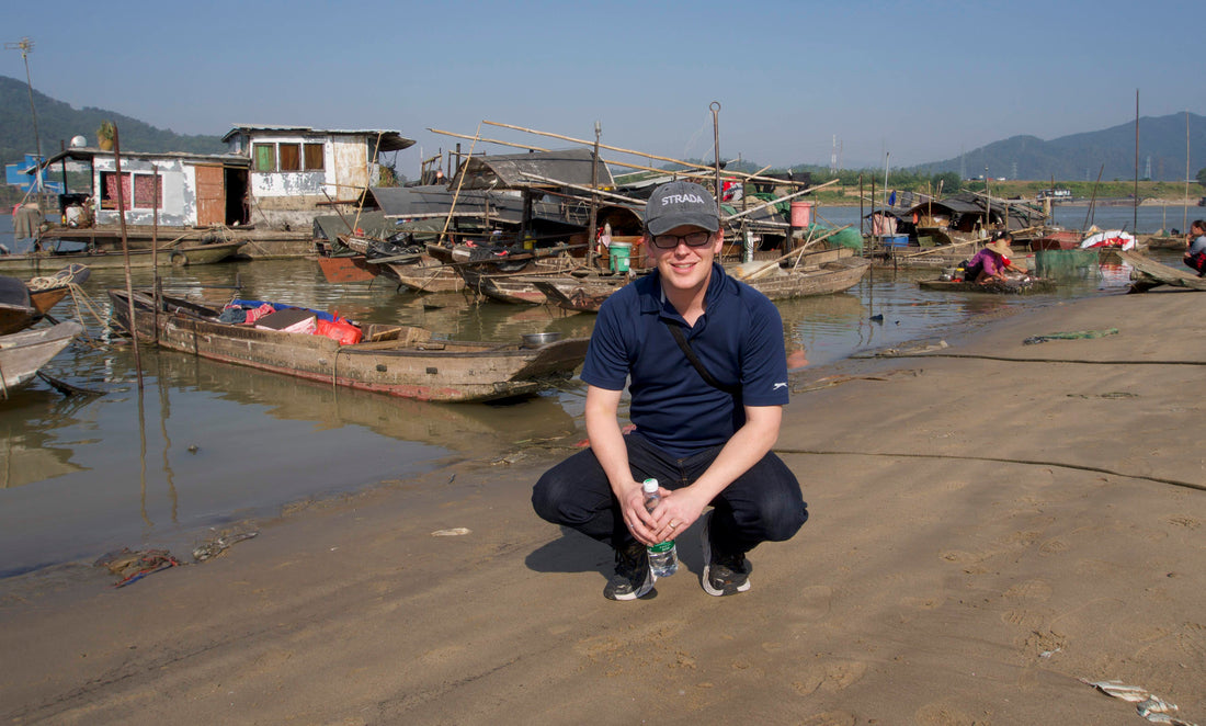 Artist in a cap crouches outdoors near water and a distant waterfront town.