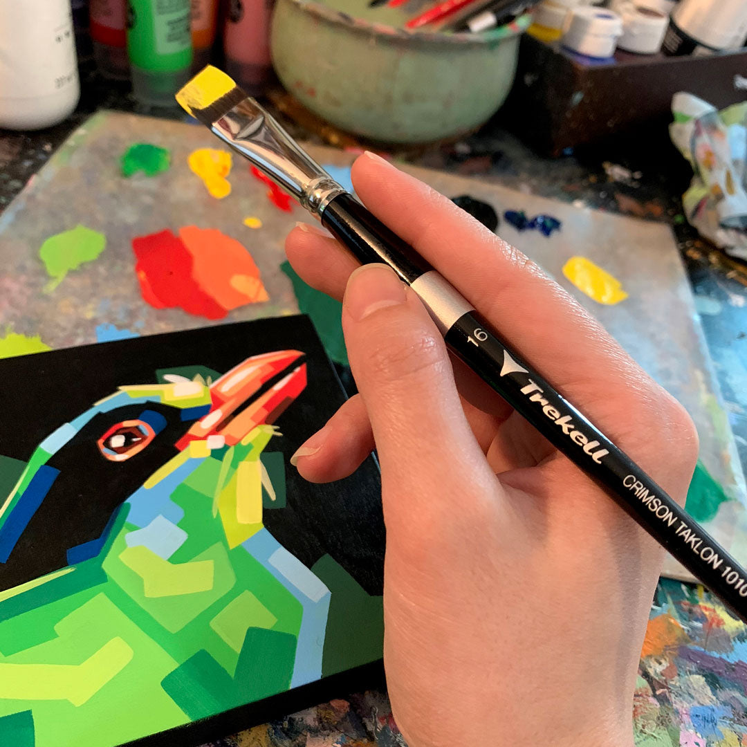 Hand paints a faceted green-and-blue bird on a small black panel.