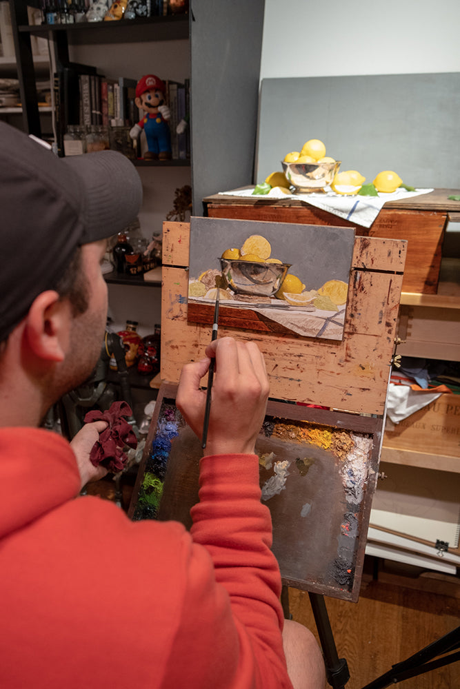 Artist in a red hoodie paints a small still life on a panel.