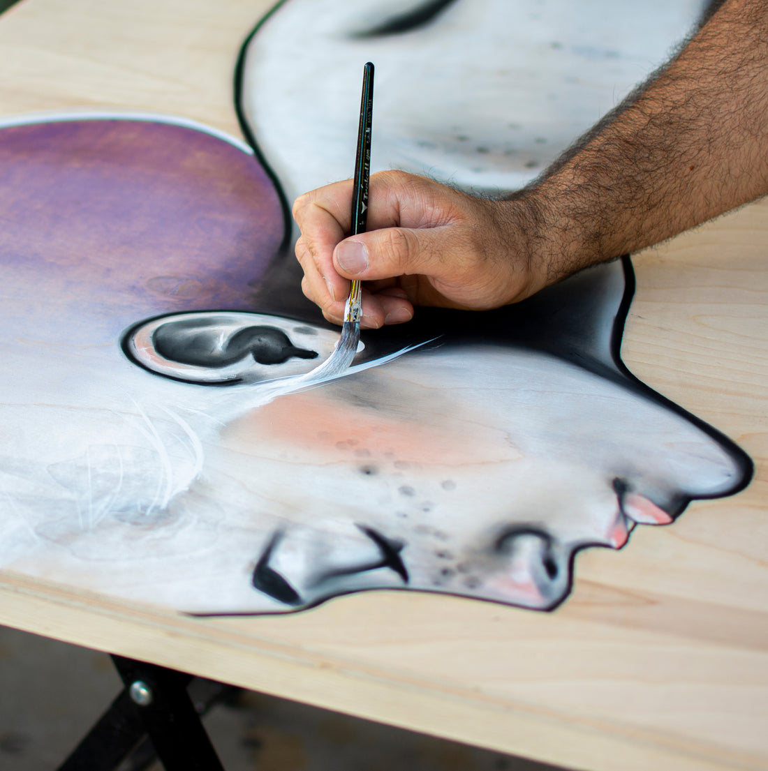Hand paints a grayscale face near the mouth on a wood panel.