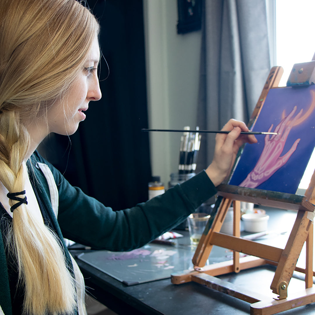 Long-haired artist paints a purple composition at a small tabletop easel.