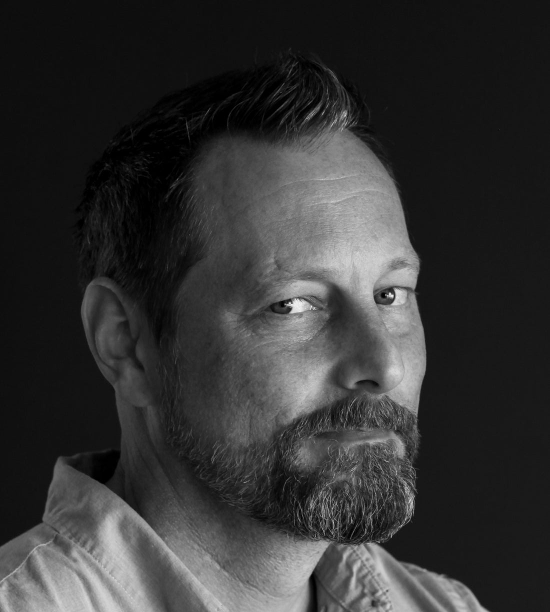 Black-and-white portrait of a short-haired man with a beard against black.
