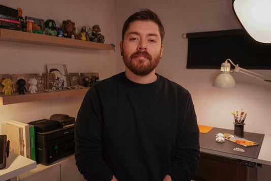 Bearded man in a black shirt stands in a studio beside shelves and a worktable.
