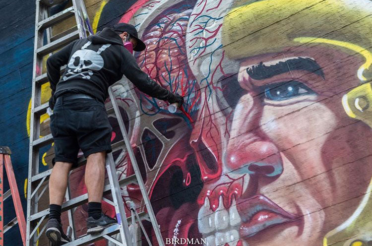 Artist on a ladder paints a large helmeted face mural.