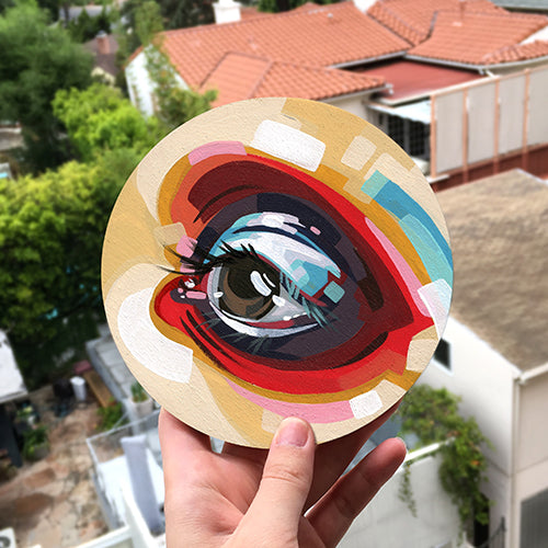 Hands hold a round panel painted with a colorful abstract eye outdoors.
