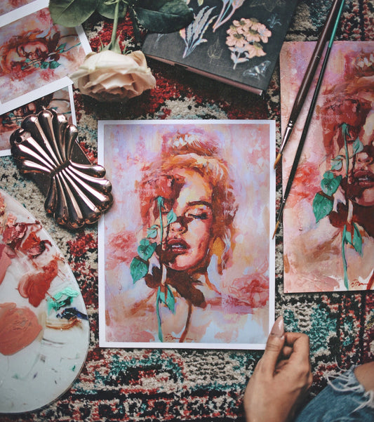 Hands hold a vivid painted portrait above a paint-covered table.