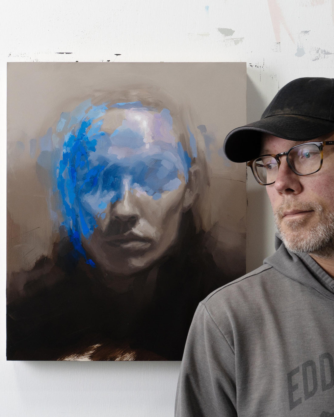 Man in a dark cap stands beside a portrait partly covered by blue paint.
