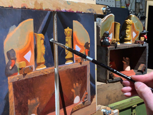 Hand paints a small still life of orange objects on a divided panel.