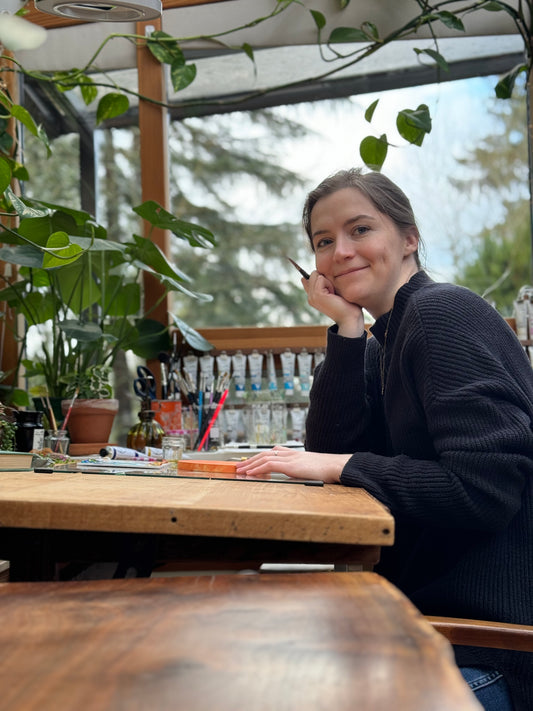 Artist sits at a studio table beneath hanging plants and large windows.