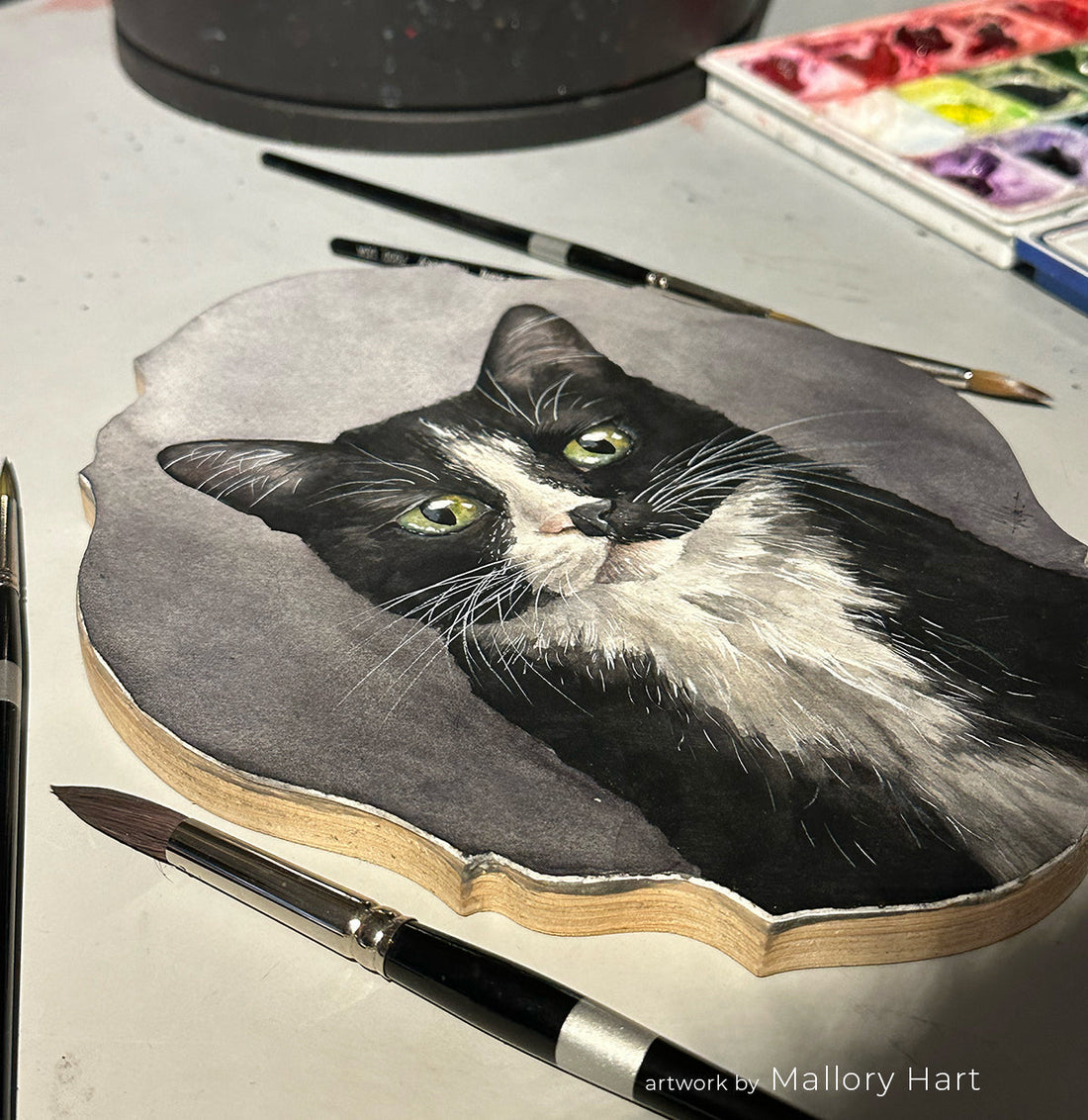 Gray-and-white cat painting beside black-handled brushes on a worktable.