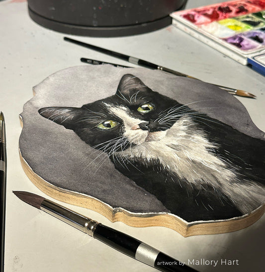 Gray-and-white cat painting beside black-handled brushes on a worktable.