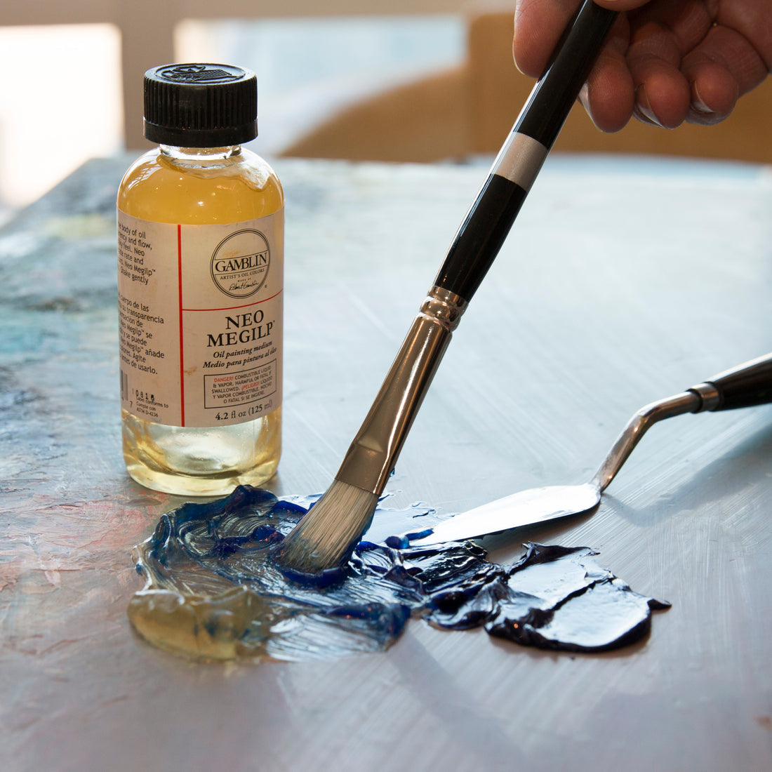 Brush spreads a pale coating beside a golden oil-medium bottle and a palette knife.