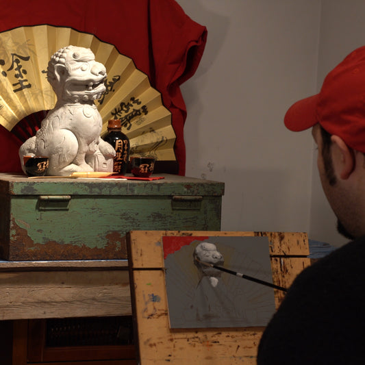 An artist in a red cap paints a still life of a white guardian-lion statue before a gold fan on a weathered green chest.