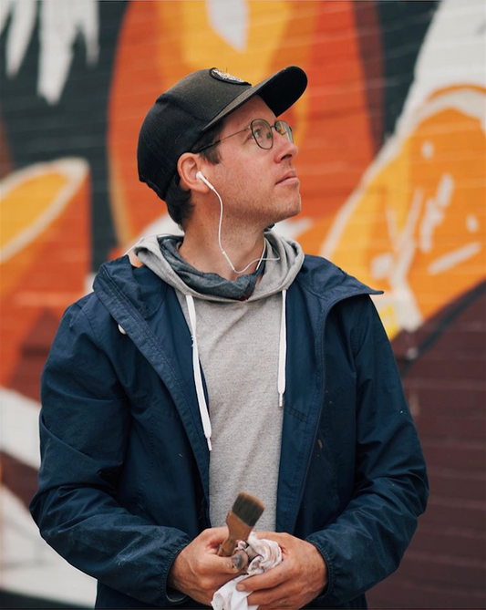 Man in a black cap and blue jacket stands before an orange mural.