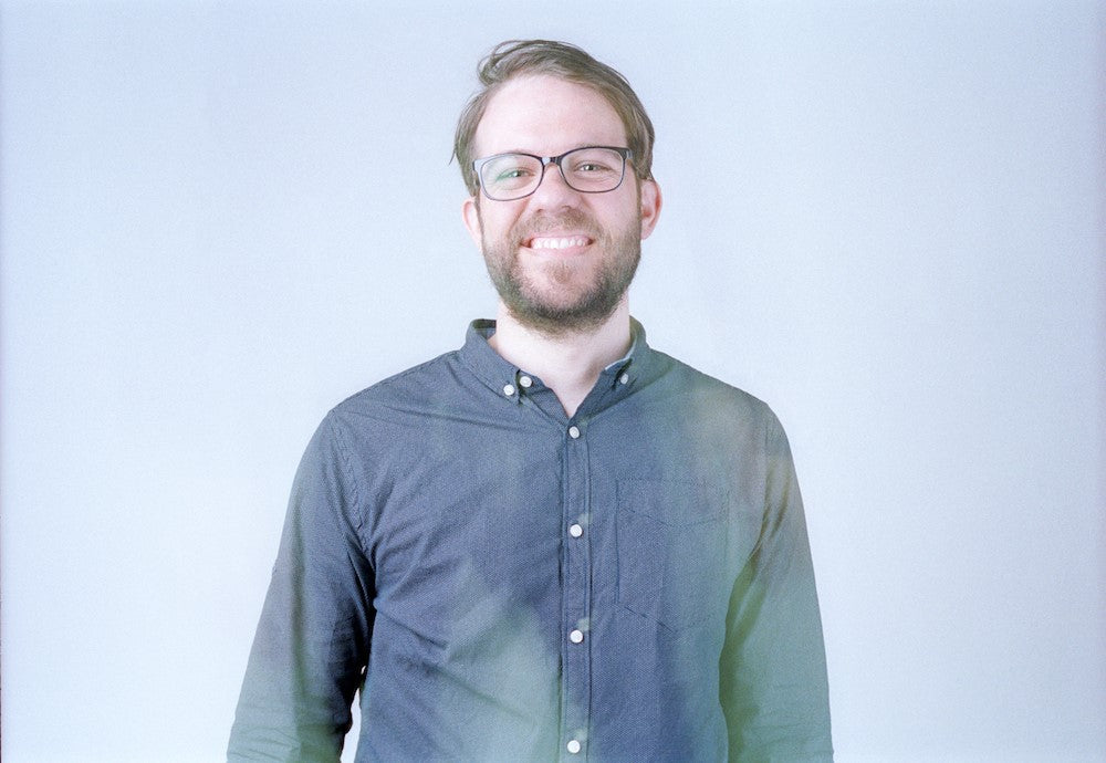 Smiling bearded man with glasses and a blue shirt against pale gray.