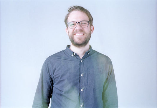 Smiling bearded man with glasses and a blue shirt against pale gray.