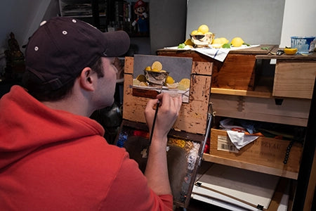 Artist in a red hoodie paints a small tabletop still life.