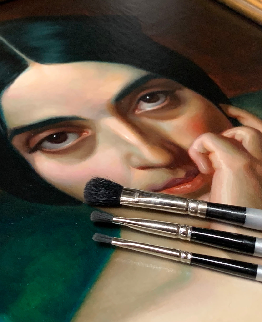 Woman holds several brushes beneath her face beside a painted portrait.