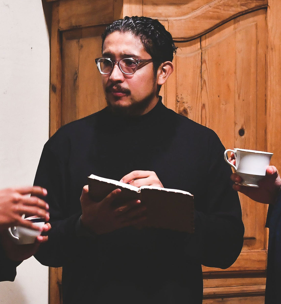 Man in dark glasses and black clothing holds a notebook before wooden doors.