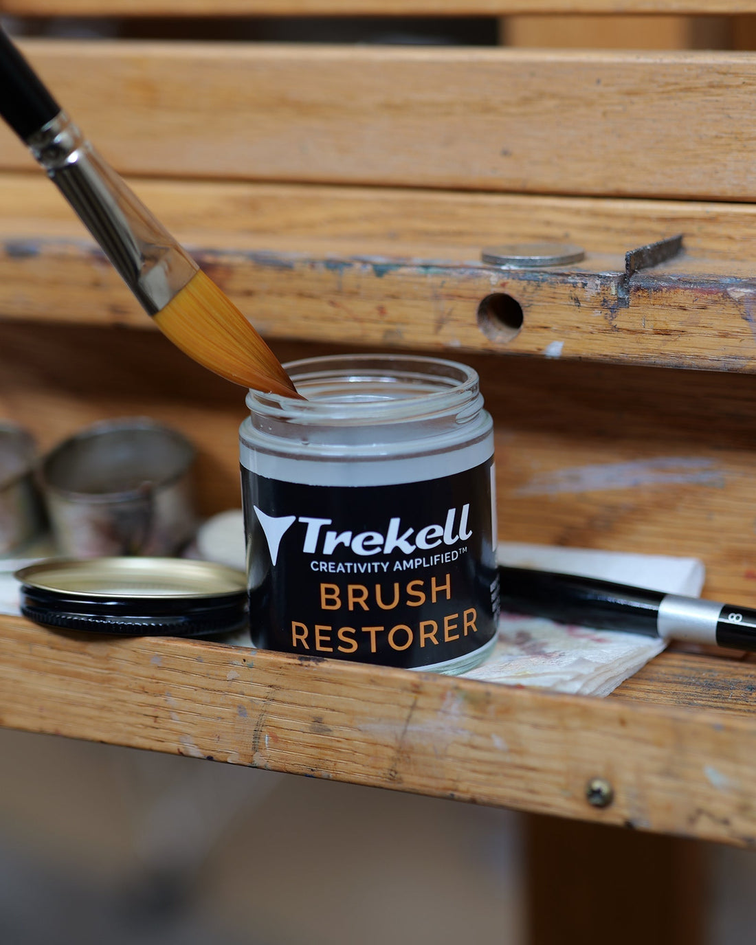 Brush dips into an open Trekell Brush Restorer jar on a wooden table.