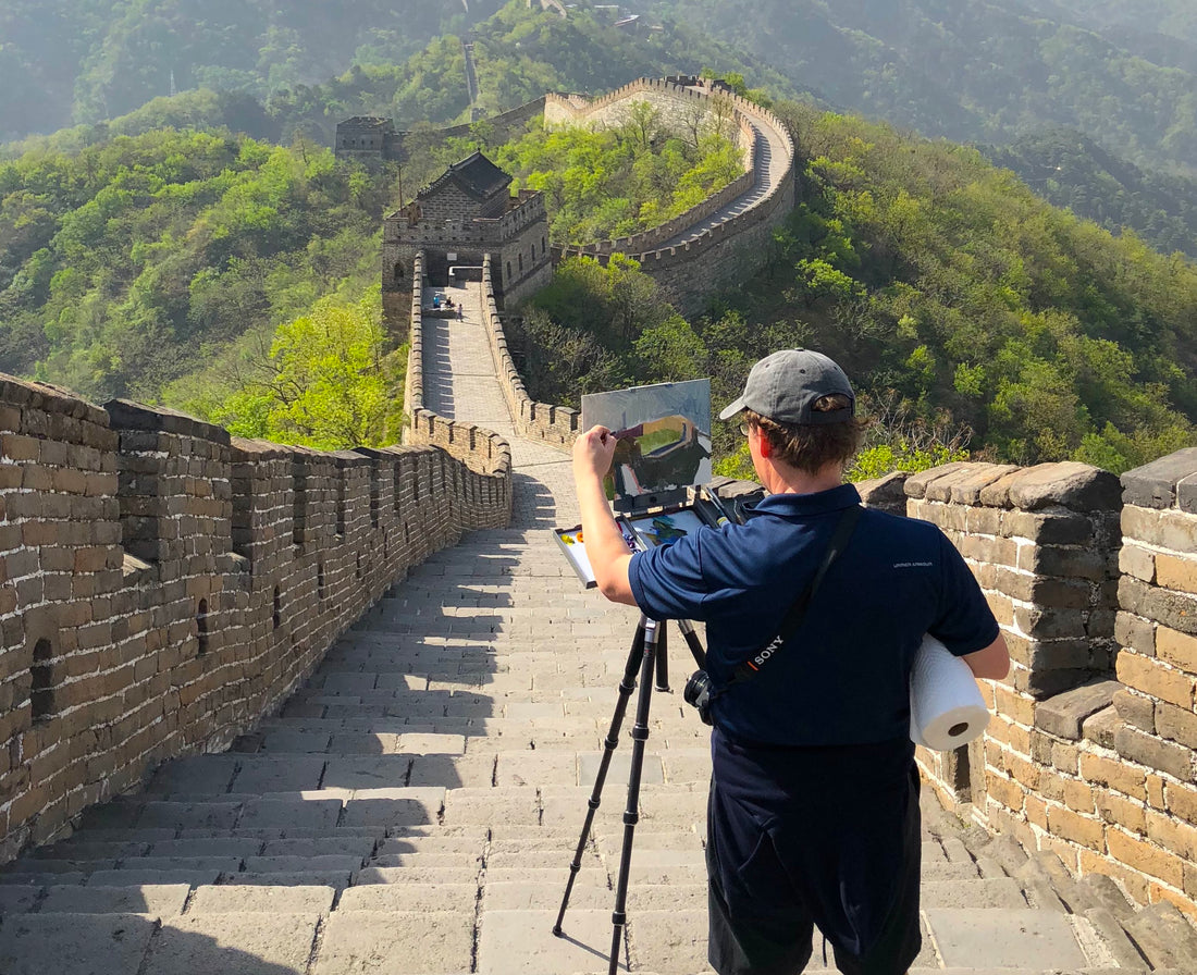 Artist paints at an easel on the Great Wall beneath a blue sky.