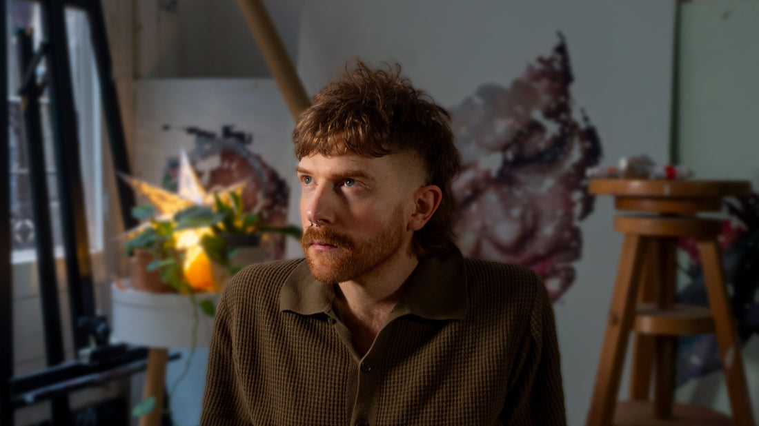 Man with short curly hair and facial hair sits in a studio with paintings.