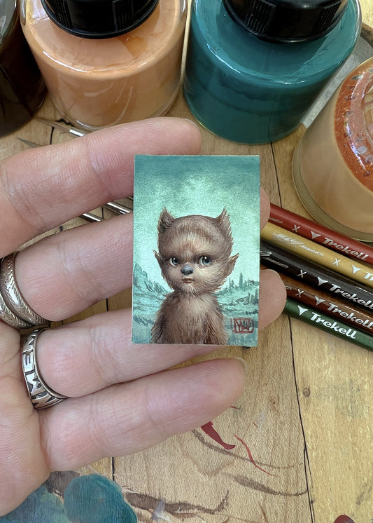 Hand holds a tiny cat portrait beside brushes and open paint containers.