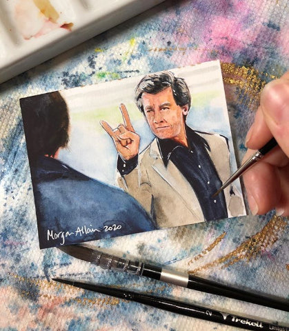 Hand holds a tiny portrait of a man in a dark suit beside brushes.