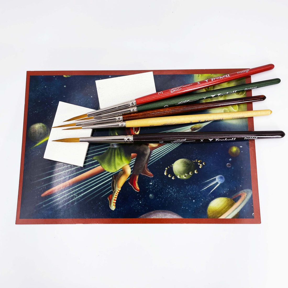 Several brushes rest across a small colorful painting with a red border.