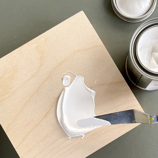 Palette knife spreads white primer on a bare square wood panel.