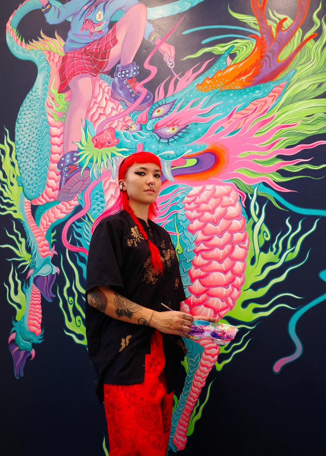 Artist in sunglasses stands before a brightly colored creature mural.