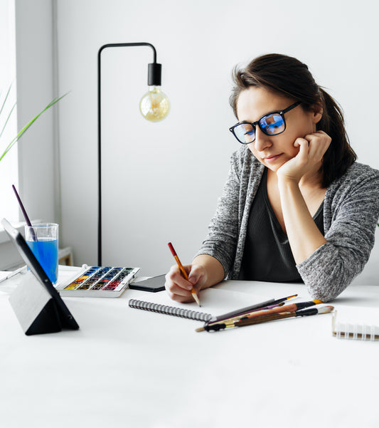 Woman with glasses writes beside a laptop at a white desk.