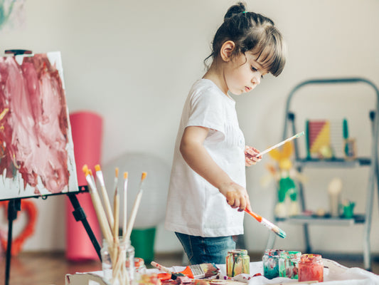 Woman paints at a table beside an easel and colorful supplies.