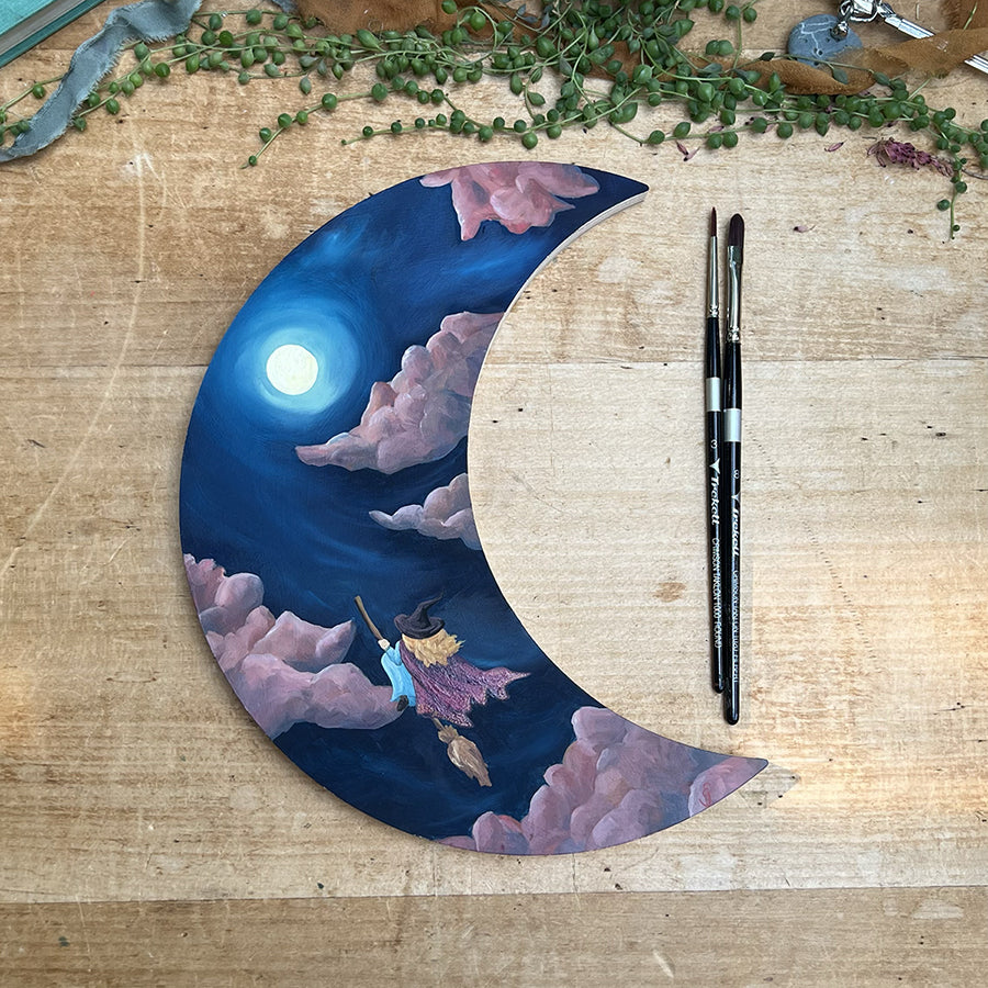 Purple crescent panel painted with clouds and a glowing moon, with two brushes lying across the open side.