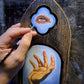 Hand paints an eye on blue above a golden hand on a black-and-gold Trekell Gothic Vienna Floater Panel.