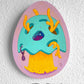 Turquoise creature with orange antlers and a yellow beard painted on a pink egg-shaped panel.