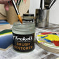 Brush resting in an open Trekell Brush Restorer jar beside a colorful paint palette.