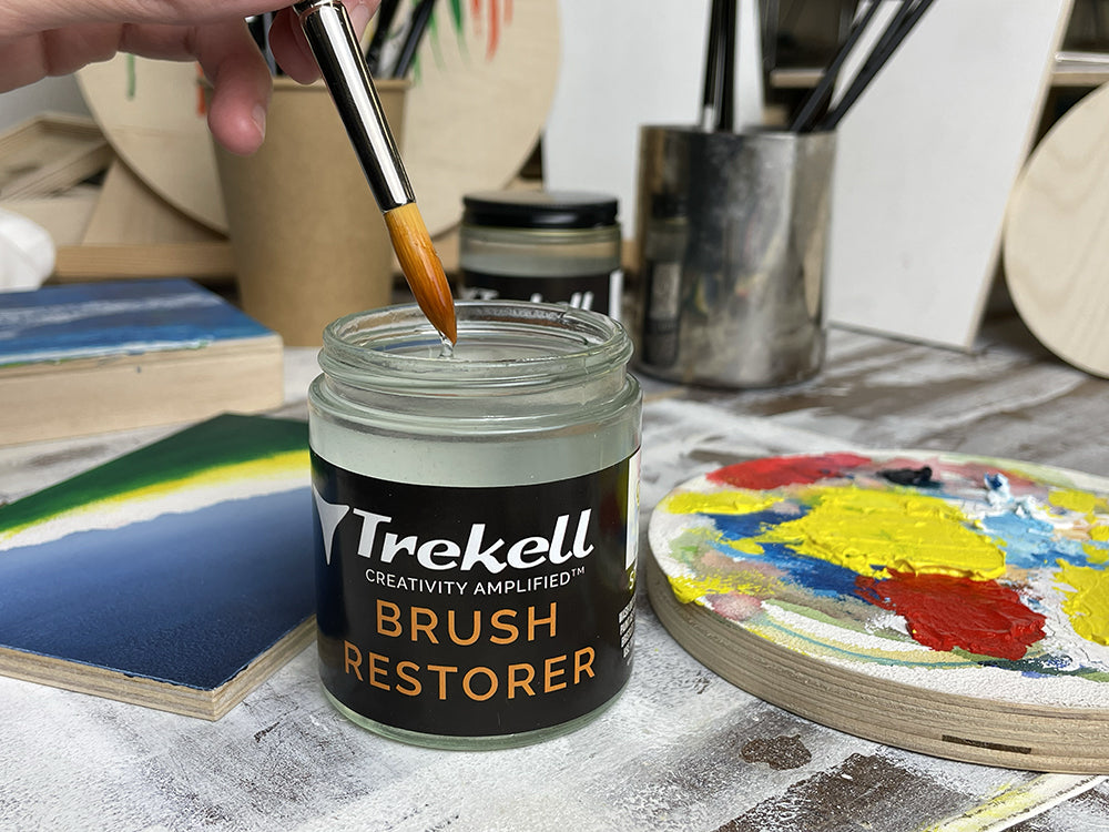 Brush resting in an open Trekell Brush Restorer jar beside a colorful paint palette.