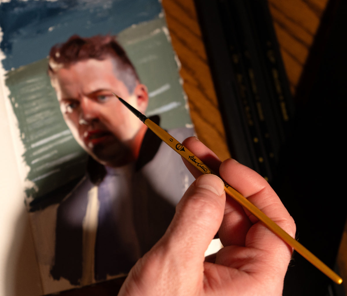 Hand holds a fine brush beside a painted portrait of a bearded man.